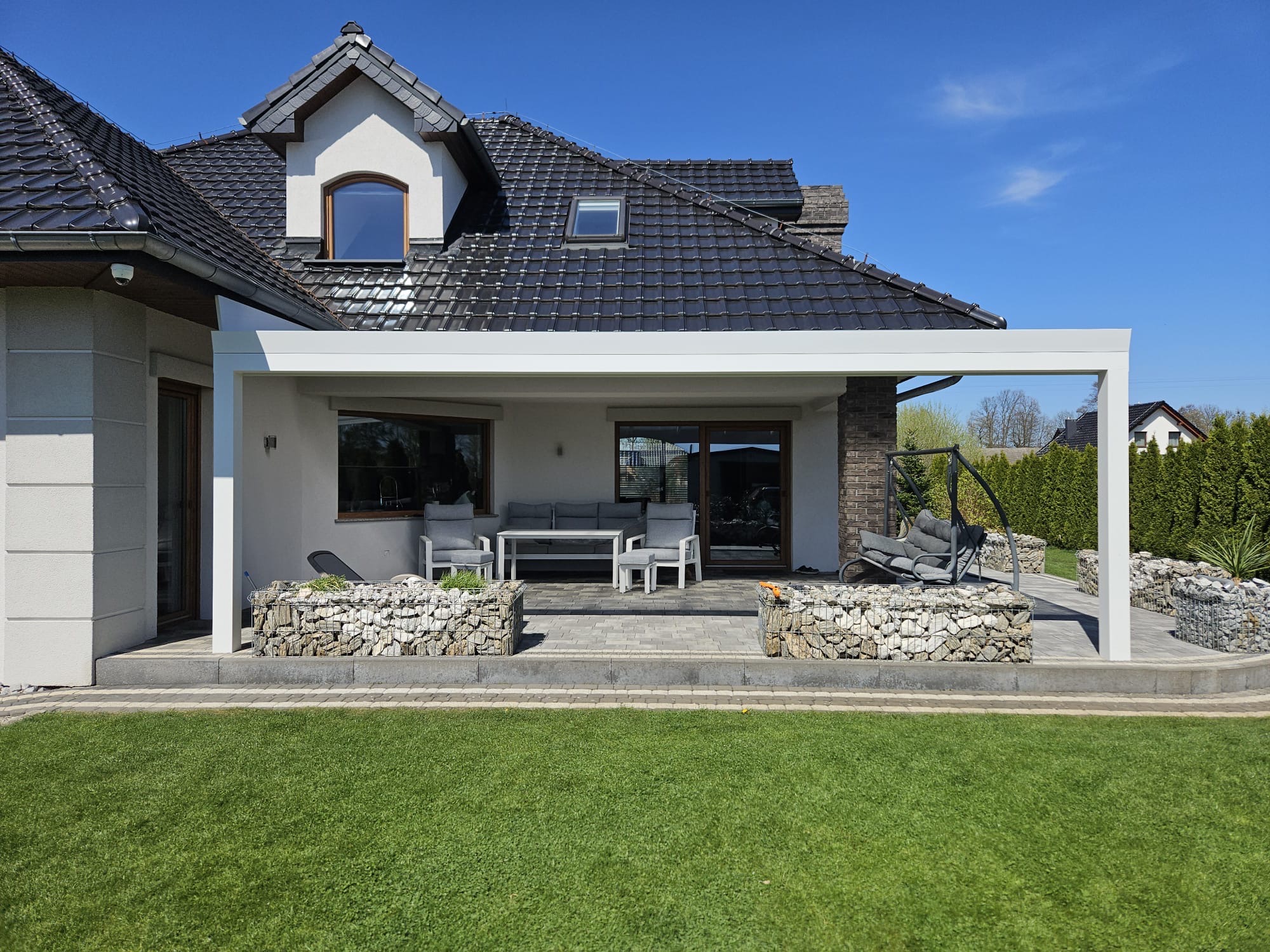 Biała, nowoczesna pergola przy domu z gabionowymi donicami i meblami ogrodowymi. Widok na zadbany trawnik i błękitne niebo. Eleganckie wykończenie tarasu.