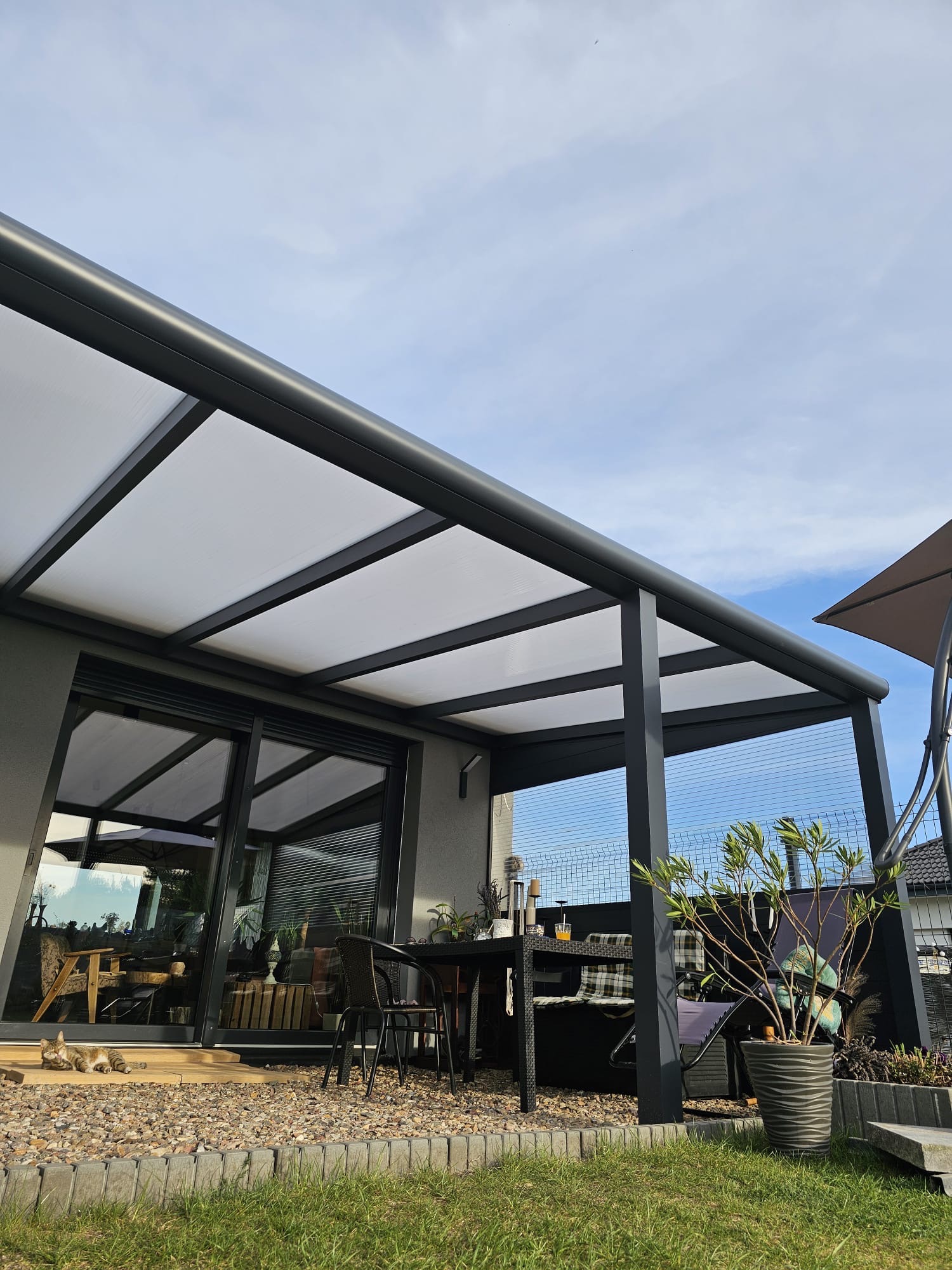 Nowoczesna, szara pergola z białym zadaszeniem, widok z dołu. Na tarasie stolik z krzesłami, roślina w donicy. W tle błękitne niebo.