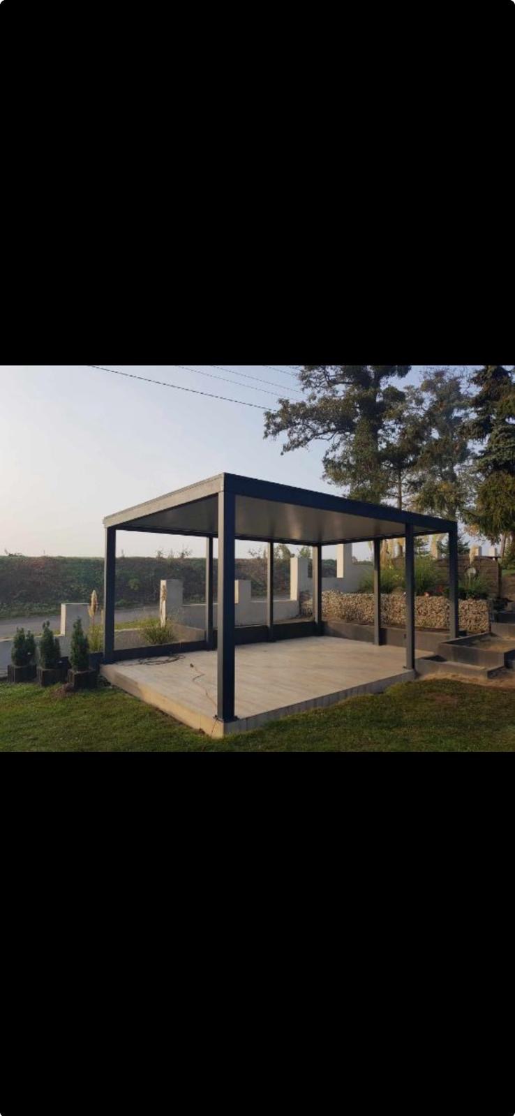 Nowoczesna, czarna pergola metalowa na betonowej platformie, otoczona zielenią i elementami dekoracyjnymi ogrodu, w słoneczny dzień.