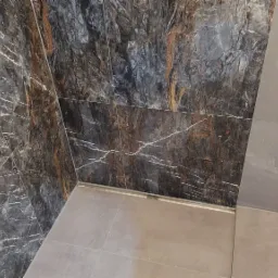 Wykończona łazienka z ciemnymi, marmurkowymi płytkami na ścianach i jasnymi płytkami na podłodze, widoczny odpływ liniowy i fragment toalety.