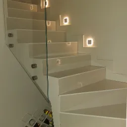 Nowoczesne schody z białymi stopniami i szklaną balustradą, podświetlane kwadratowymi lampami w ścianie, z półką na wino pod pierwszym stopniem.