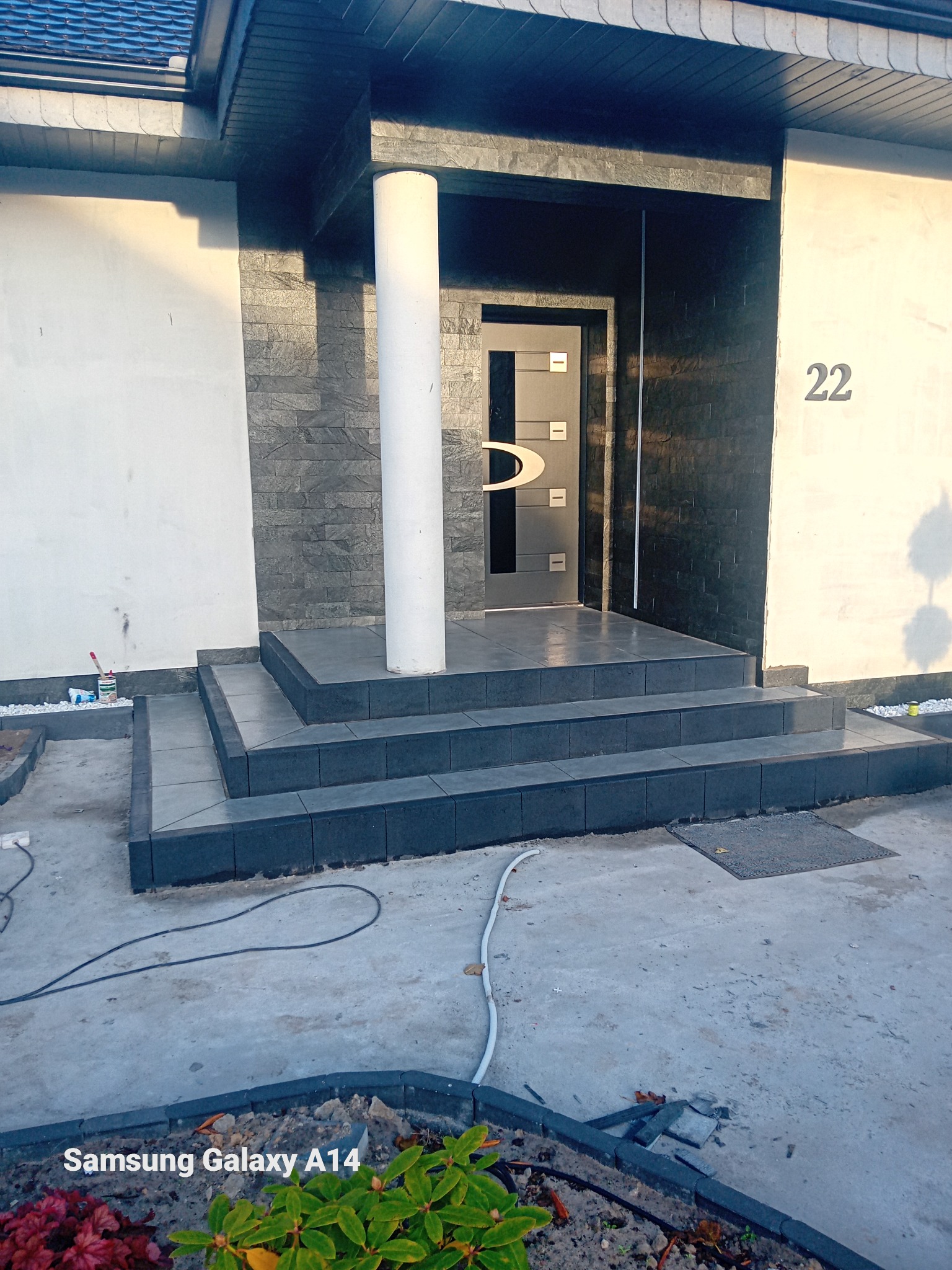 Wejście do domu z ciemnoszarą elewacją imitującą kamień, białą kolumną i schodami z ciemnych płytek, numer domu 22 widoczny na białej ścianie, w tle widoczne rośliny.