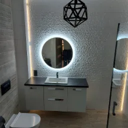 Nowoczesna łazienka z umywalką na szafce, okrągłym lustrem z podświetleniem LED, geometryczną lampą wiszącą i trójwymiarowymi płytkami ściennymi.