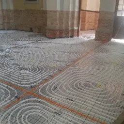 Instalacja ogrzewania podłogowego w trakcie montażu w pomieszczeniu z surowymi ścianami z cegły i kolumnami, widoczne białe rury ułożone spiralnie na siatce montażowej.