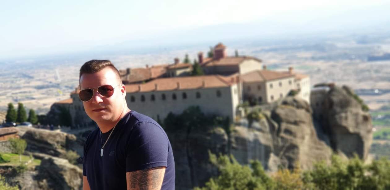 Mężczyzna w okularach przeciwsłonecznych pozuje na tle malowniczego klasztoru Meteora w Grecji. W tle rozległy krajobraz.