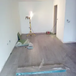 Wnętrze pokoju w trakcie układania paneli podłogowych, widoczne paczki paneli Krono Original, lampa budowlana na trójnogu i częściowo ułożona podłoga o szarym odcieniu.