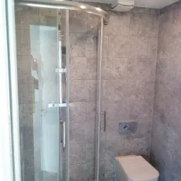 Nowoczesna kabina prysznicowa z deszczownicą i szklanymi drzwiami, obok toaleta w szaro-białej łazience wykończonej płytkami imitującymi kamień.