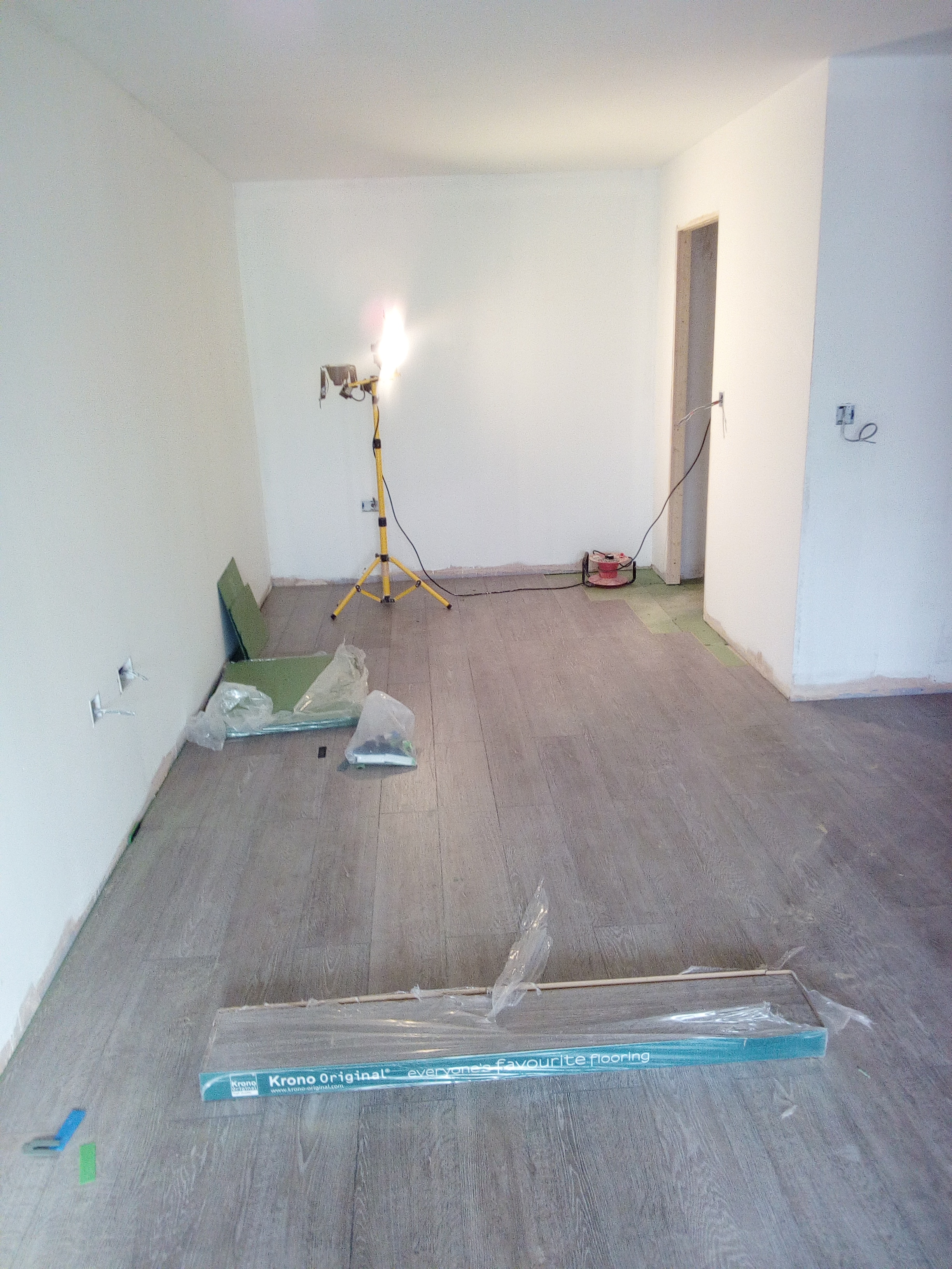 Wnętrze pokoju w trakcie układania paneli podłogowych, widoczne paczki paneli Krono Original, lampa budowlana na trójnogu i częściowo ułożona podłoga o szarym odcieniu.
