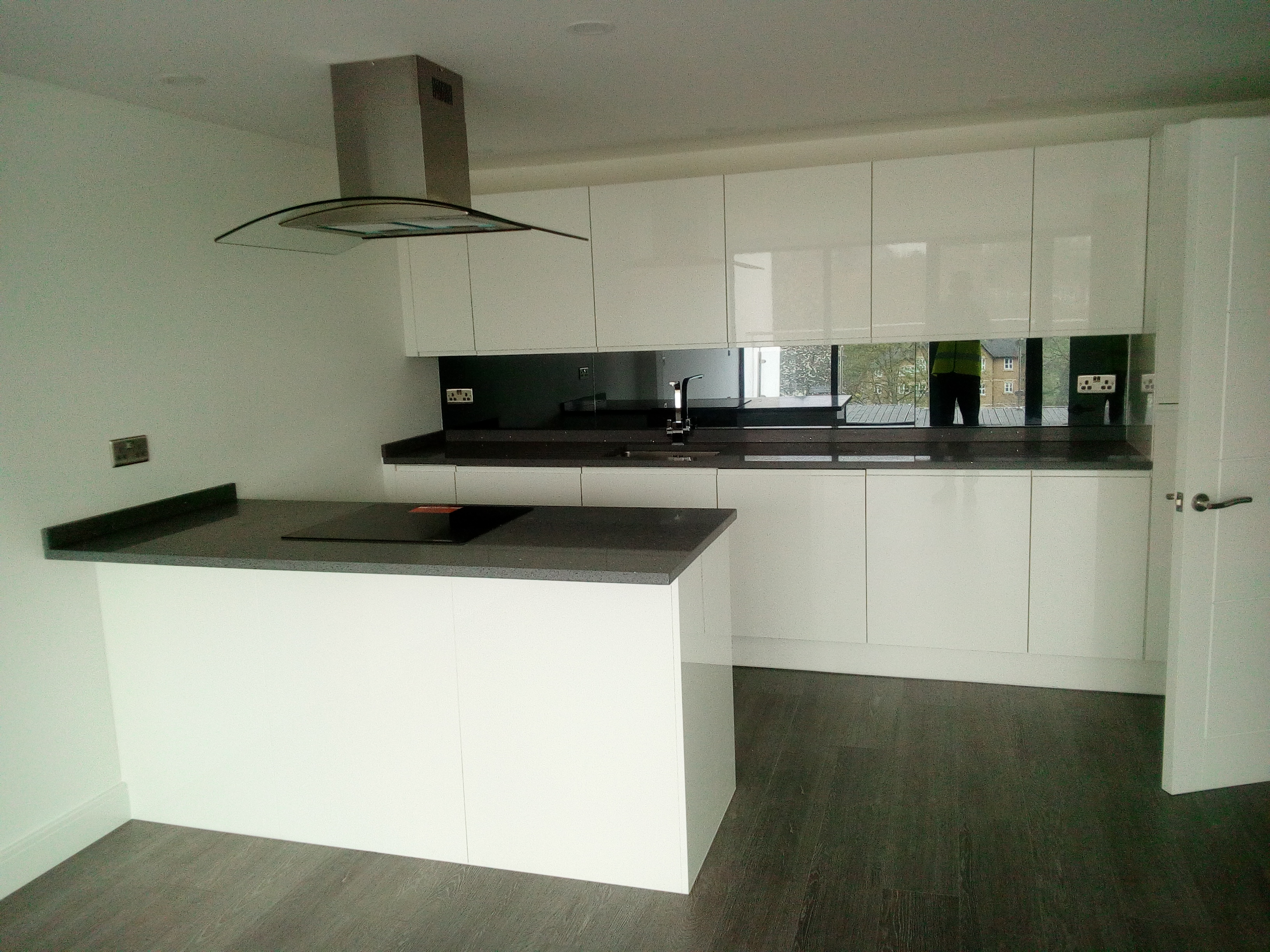 Nowoczesna, minimalistyczna kuchnia z białymi, błyszczącymi szafkami, szarym blatem i płytą indukcyjną na wyspie. W tle widoczne okno z odbiciem sylwetki osoby oraz ciemna szklana ściana nad blatem.