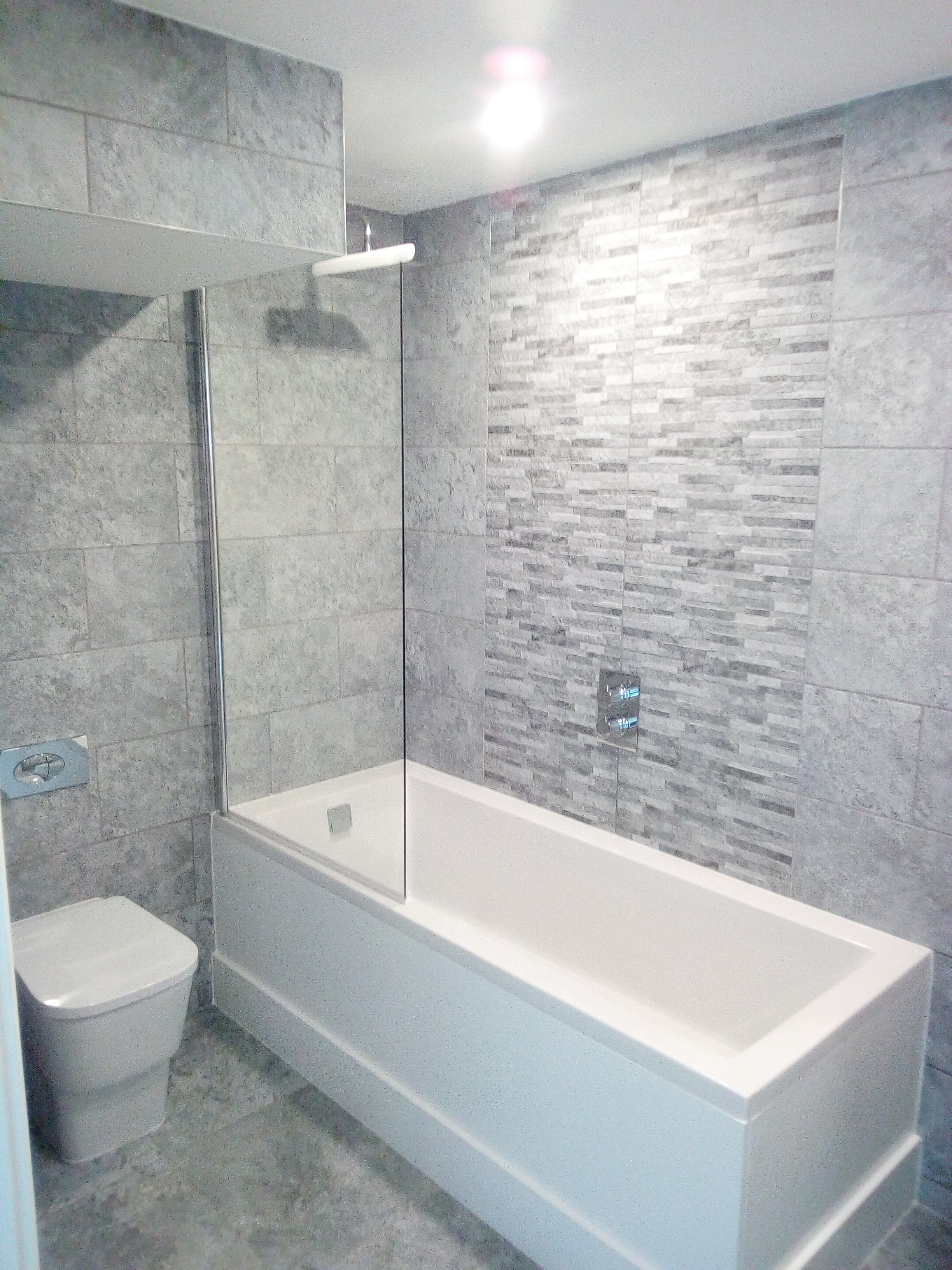 Wykończona łazienka w odcieniach szarości z wanną z parawanem, toaletą i mozaiką na ścianie, oświetlona górnym światłem.