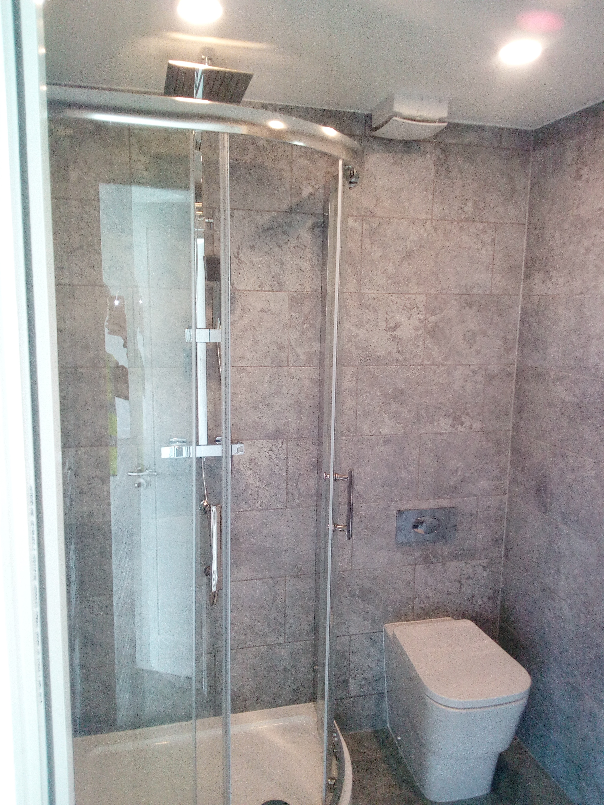 Nowoczesna kabina prysznicowa z deszczownicą i szklanymi drzwiami, obok toaleta w szaro-białej łazience wykończonej płytkami imitującymi kamień.