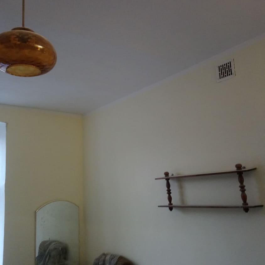 Wnętrze pokoju z beżowymi ścianami, widoczna drewniana półka na ścianie, okrągła lampa sufitowa z brązowym kloszem, lustro w złotej ramie oraz kratka wentylacyjna na suficie.