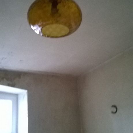 Pokój przed remontem: surowy tynk na ścianach, widoczny narożnik, okno i wisząca lampa z żółtym kloszem.