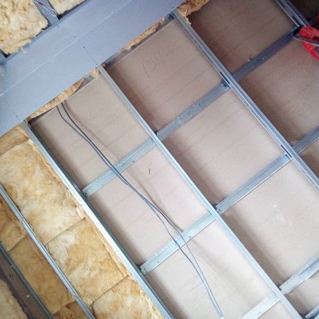 Widok z dołu na metalową konstrukcję sufitu podwieszanego z zamontowaną płytą i izolacją z wełny mineralnej, widoczne przewody elektryczne.