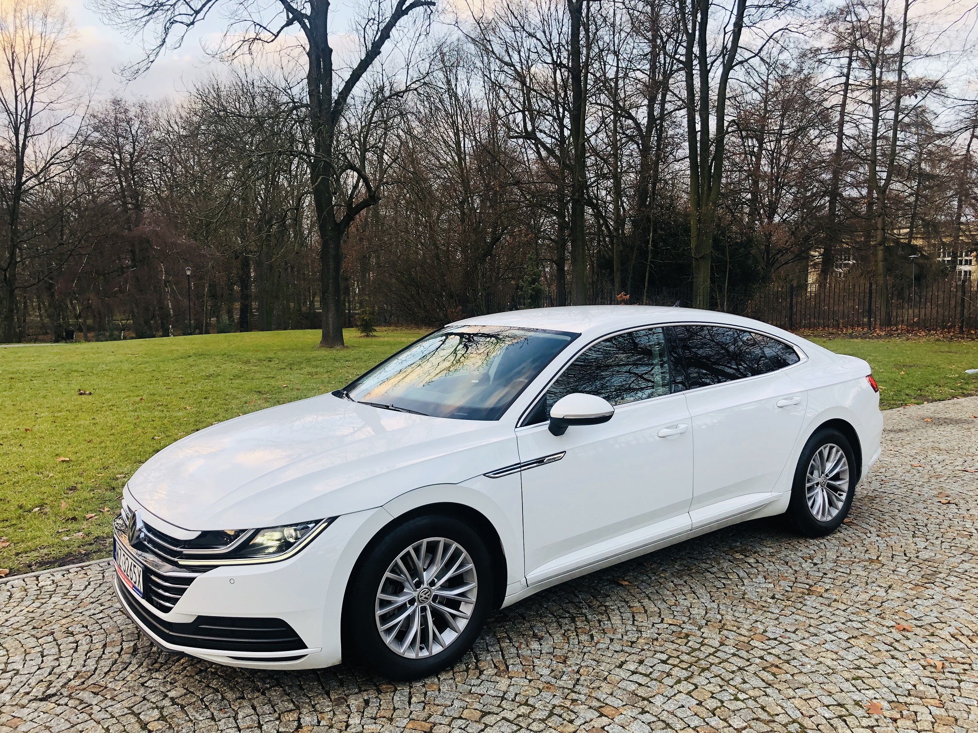 Biały samochód osobowy marki Volkswagen Arteon, zaparkowany na brukowanej nawierzchni, z drzewami w tle.