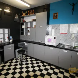 Szare szafki i blat z czarnym wykończeniem w pomieszczeniu z czarno-białą podłogą w szachownicę; na blacie sterylizator i umywalka; na ścianie grafiki i instrukcje higieny.