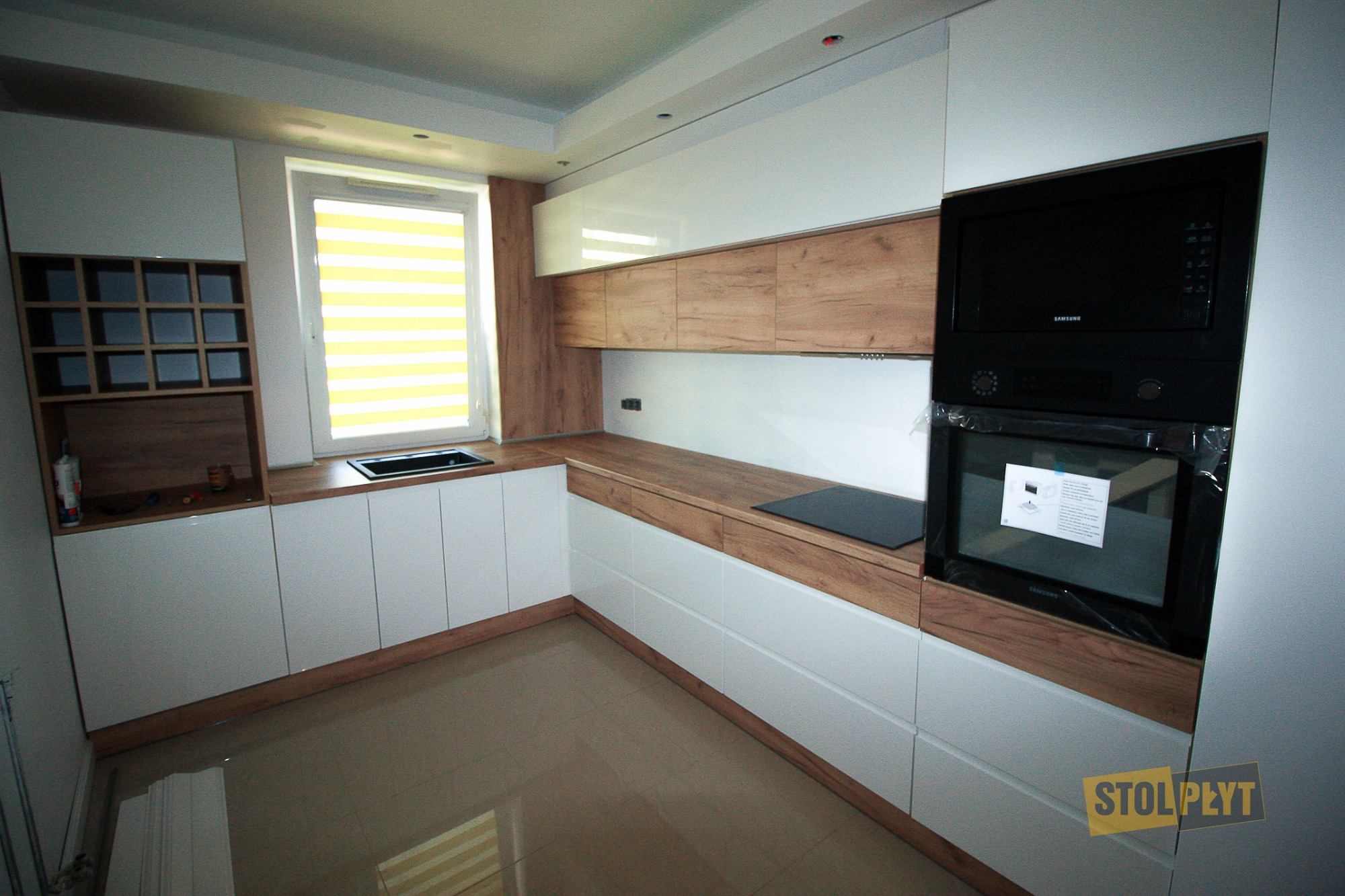 Nowoczesna kuchnia z białymi frontami i drewnianymi blatami, z piekarnikiem Samsung jeszcze w folii ochronnej, widok na okno z żaluzjami w kolorze żółtym i brązowe płytki na podłodze.