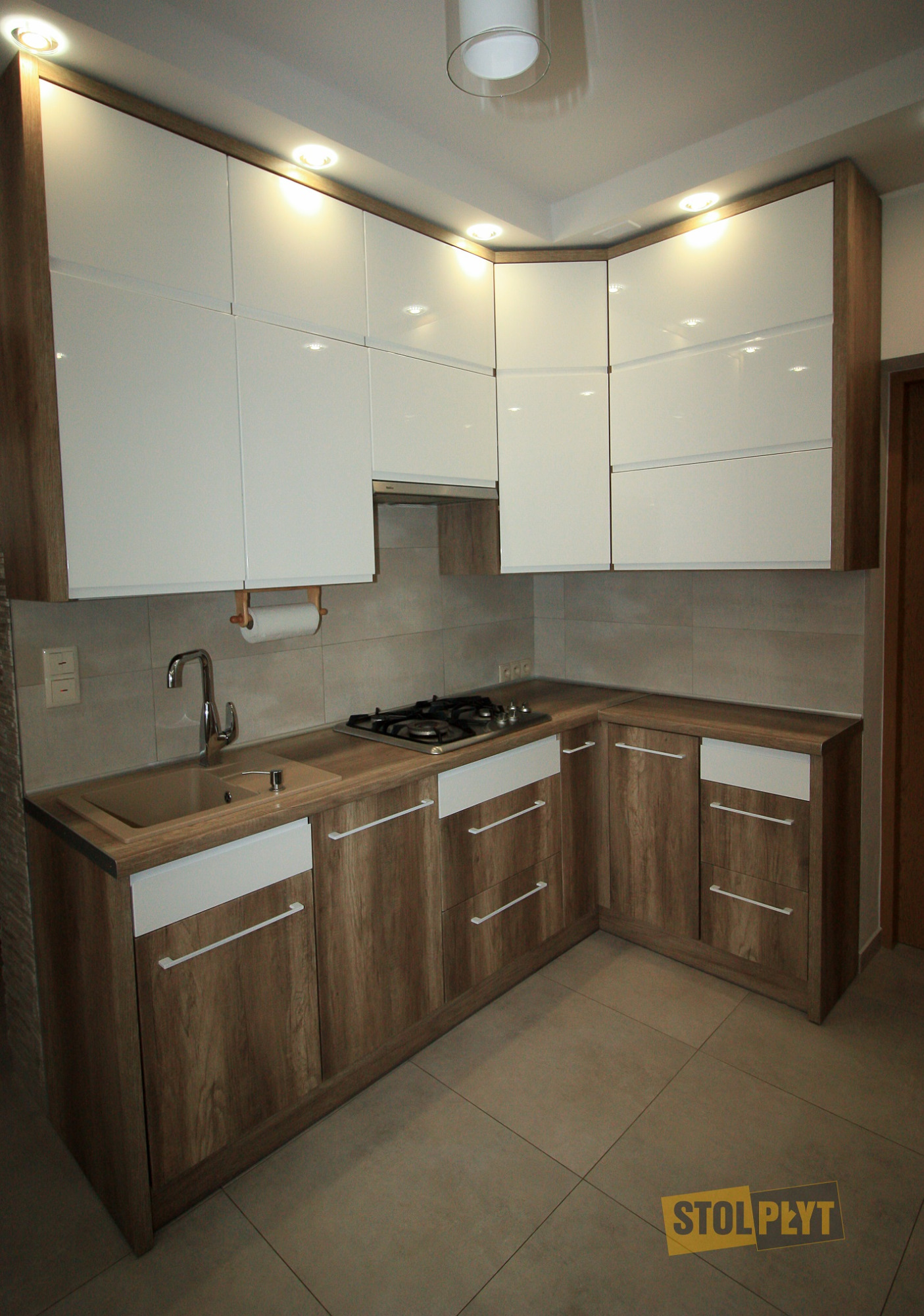 Kremowo-biała kuchnia narożna na wymiar z drewnopodobnymi frontami dolnych szafek i białymi górnymi, oświetlona halogenami, z beżowym blatem i płytkami w tle.