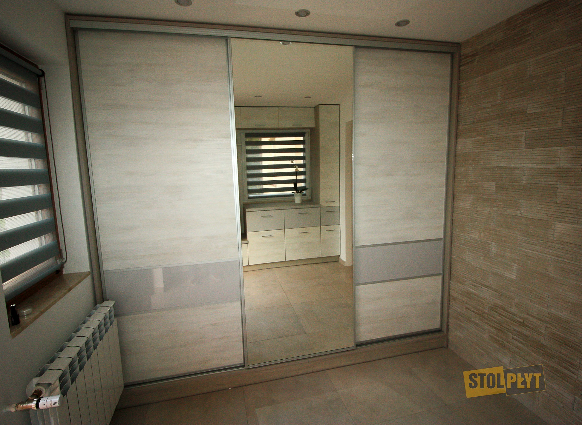 Wnętrze z przesuwnymi drzwiami szafy wnękowej, widok na jasną kuchnię z żaluzjami w oknie, ściana wykończona dekoracyjną cegłą, widoczny grzejnik i logo firmy Stol Płyt.