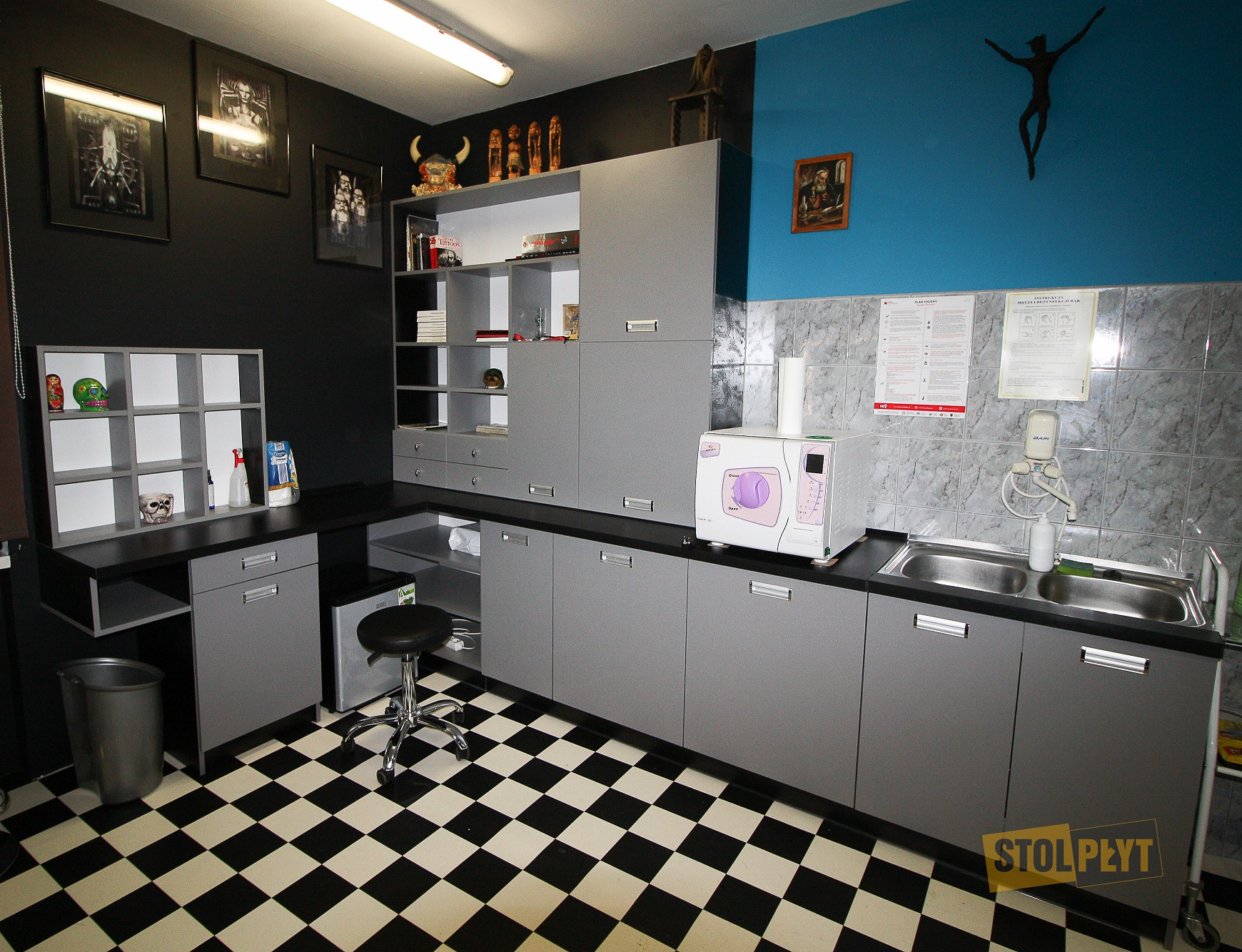 Szare szafki i blat z czarnym wykończeniem w pomieszczeniu z czarno-białą podłogą w szachownicę; na blacie sterylizator i umywalka; na ścianie grafiki i instrukcje higieny.