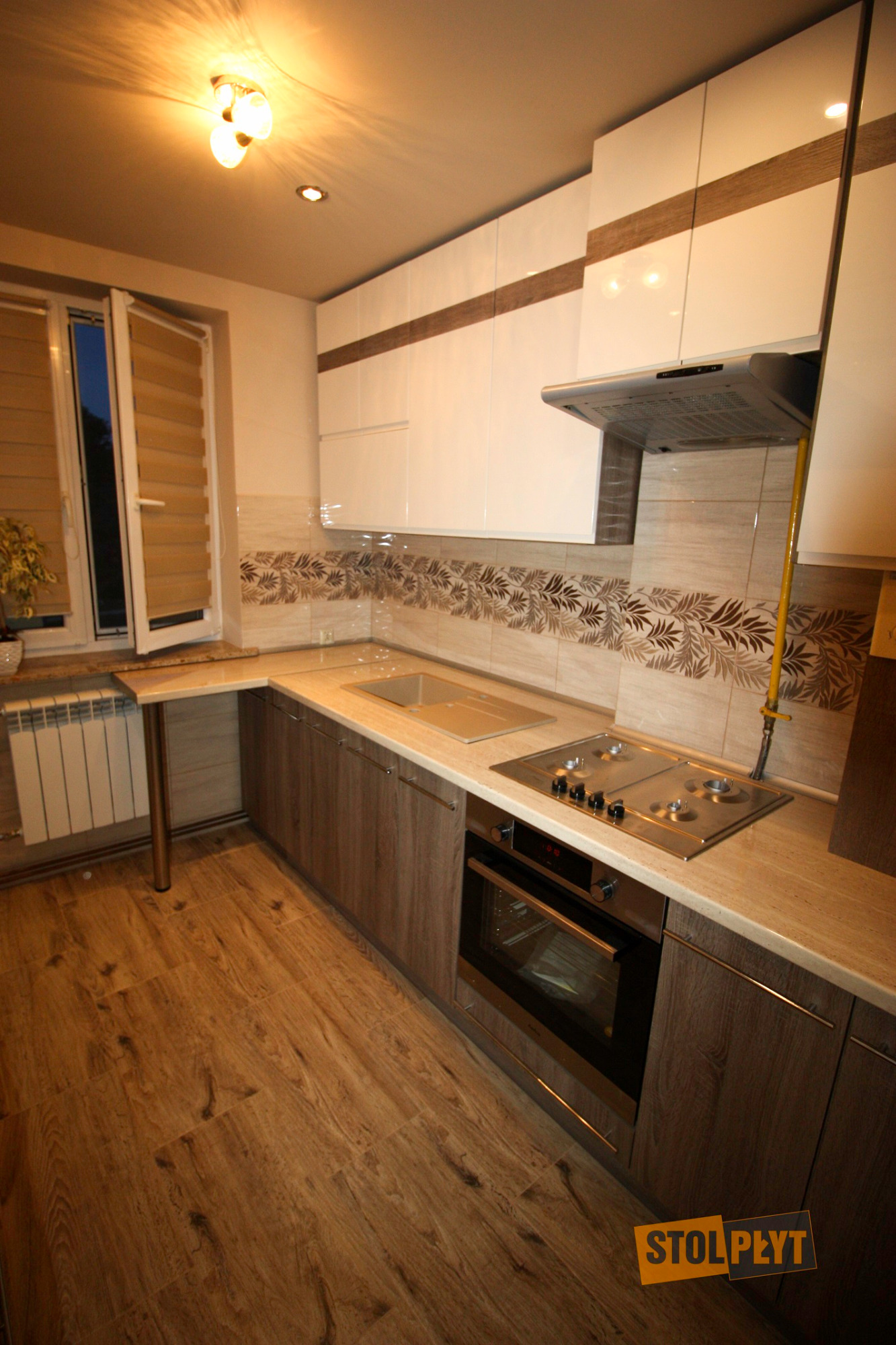 Jasna kuchnia z drewnianymi akcentami, płyta grzewcza i piekarnik w zabudowie, widok na okno z żaluzjami, drewniana podłoga, dekoracyjne płytki na ścianie