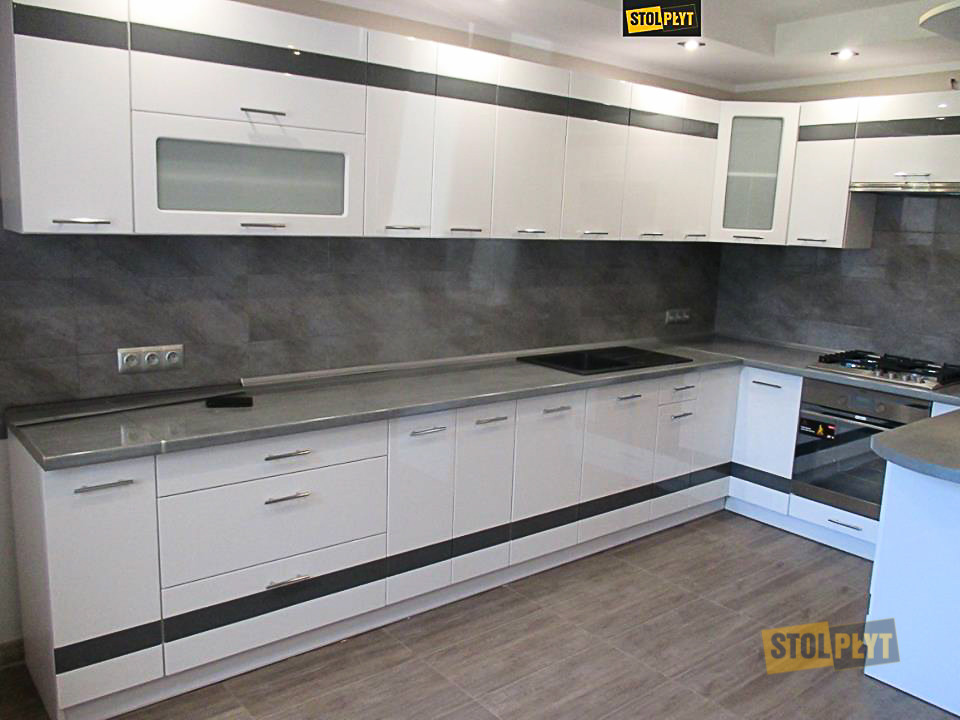 Biała kuchnia z szarym blatem i elementami dekoracyjnymi w kolorze antracytu, widok na zabudowę kuchenną z płytą grzewczą i piekarnikiem, szara ściana nad blatem.