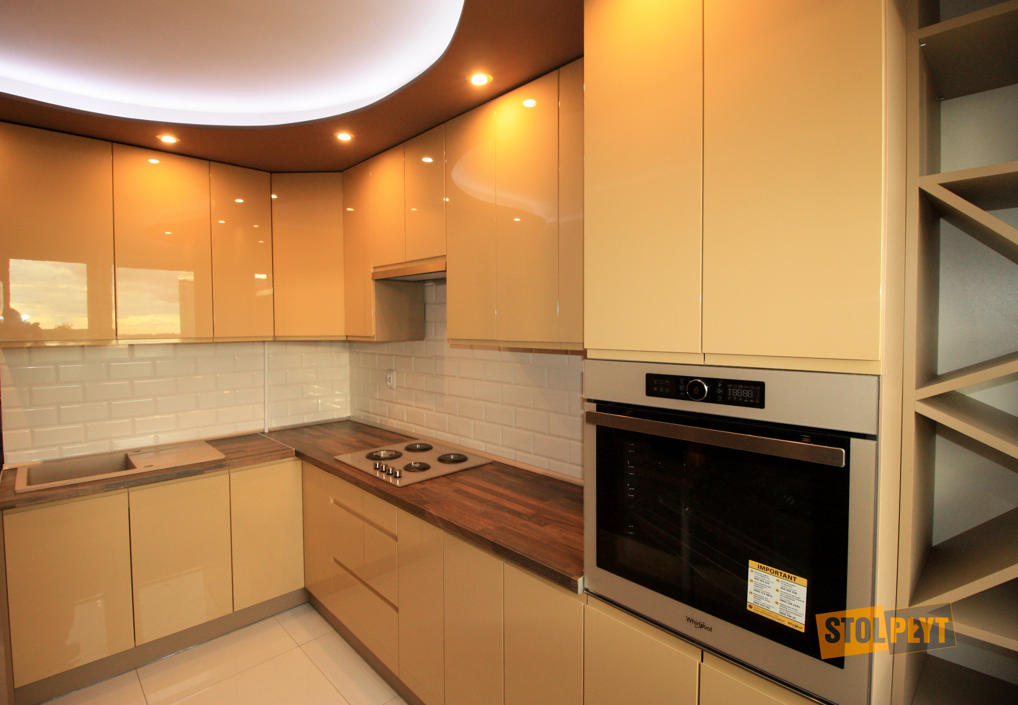 Beżowa zabudowa kuchenna z połyskiem, drewniany blat, białe płytki metra, oświetlenie punktowe i piekarnik Whirlpool z naklejką informacyjną.