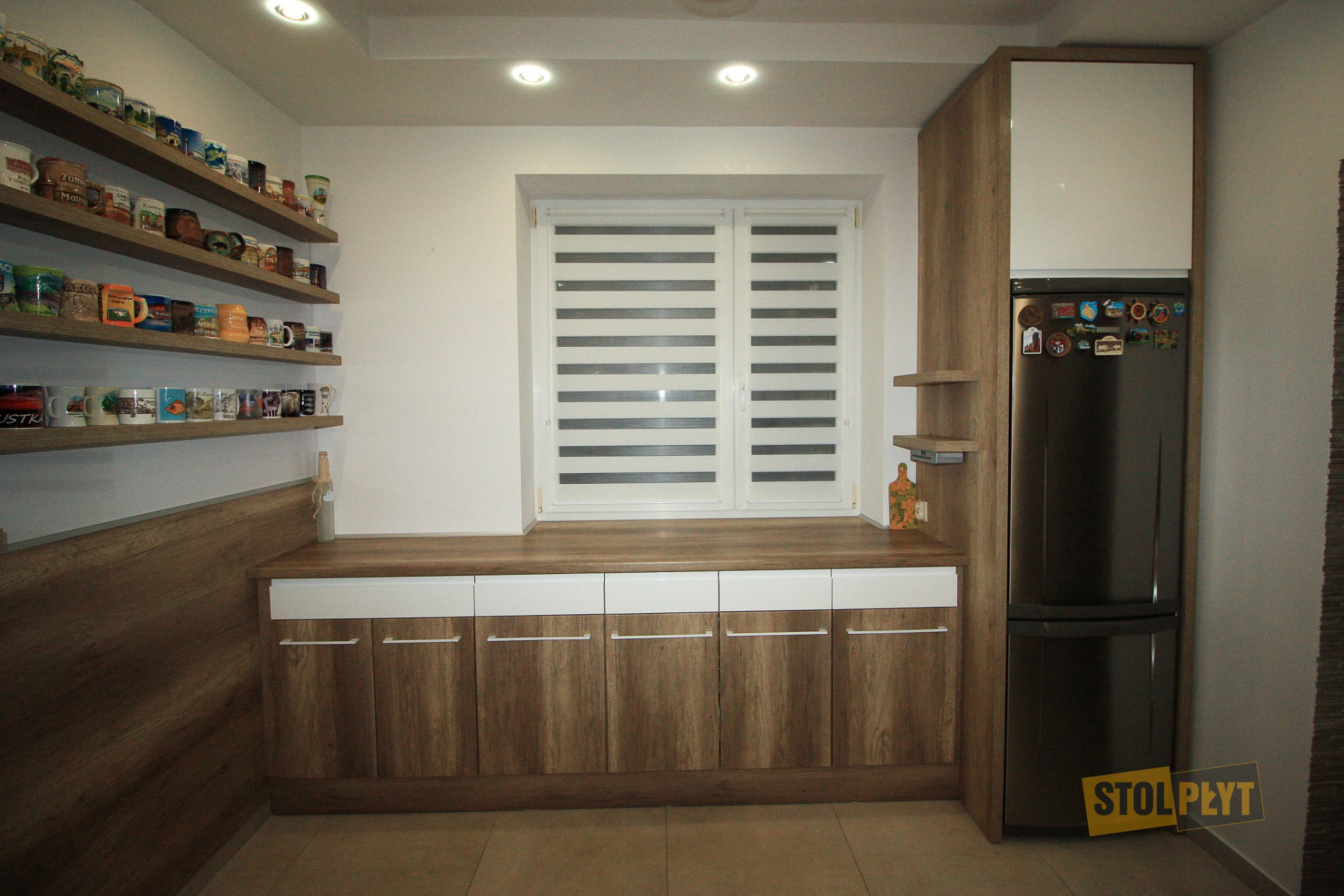 Kuchnia z drewnianymi frontami szafek dolnych z białymi szufladami, blat pod oknem, półki z kolekcją kubków, czarna lodówka z magnesami.