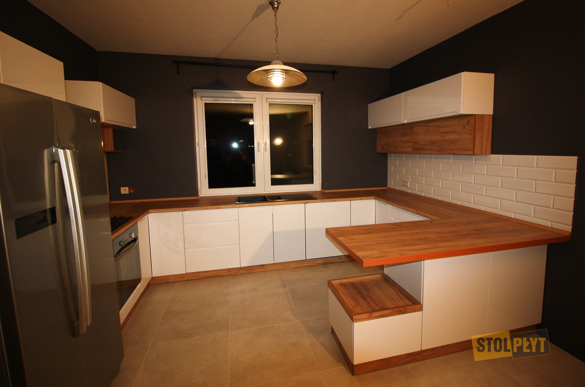 Nowoczesna kuchnia z białymi frontami szafek, drewnianym blatem i wyspą z siedziskiem, oświetlona wiszącą lampą, z widocznym fragmentem lodówki i białymi płytkami na ścianie.