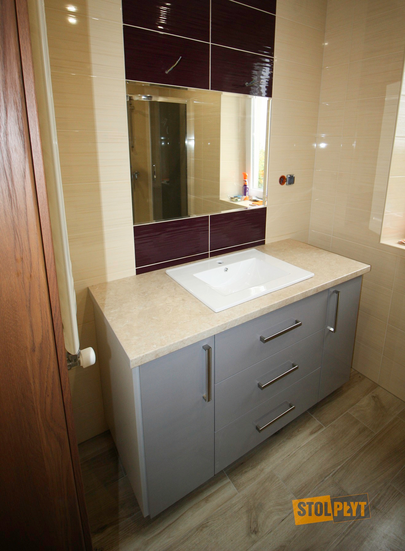 Szara szafka łazienkowa z trzema szufladami pod umywalką, beżowy blat, lustro z bordowymi akcentami, beżowe płytki na ścianach i podłodze imitujące drewno, widoczny zawór grzejnika.