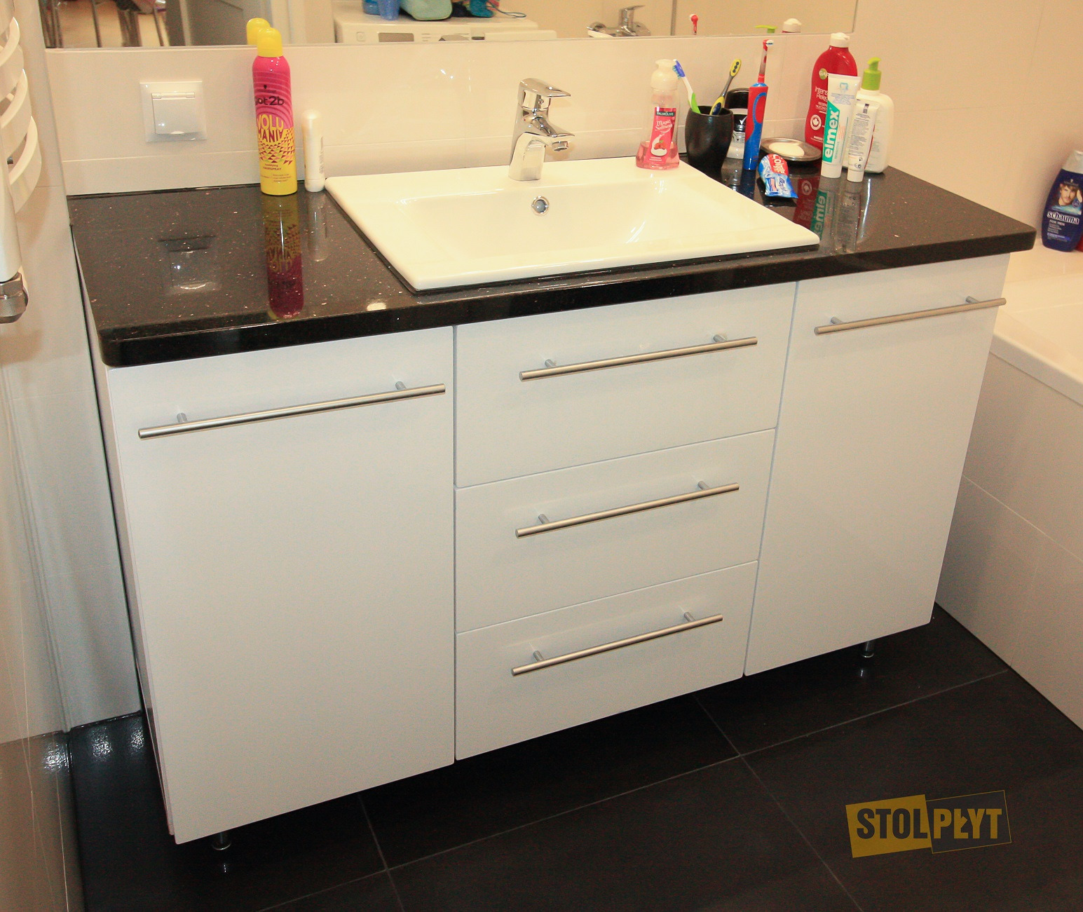 Biała szafka łazienkowa z trzema szufladami i czarnym, błyszczącym blatem pod umywalkę; widoczne srebrne uchwyty i czarne płytki podłogowe.