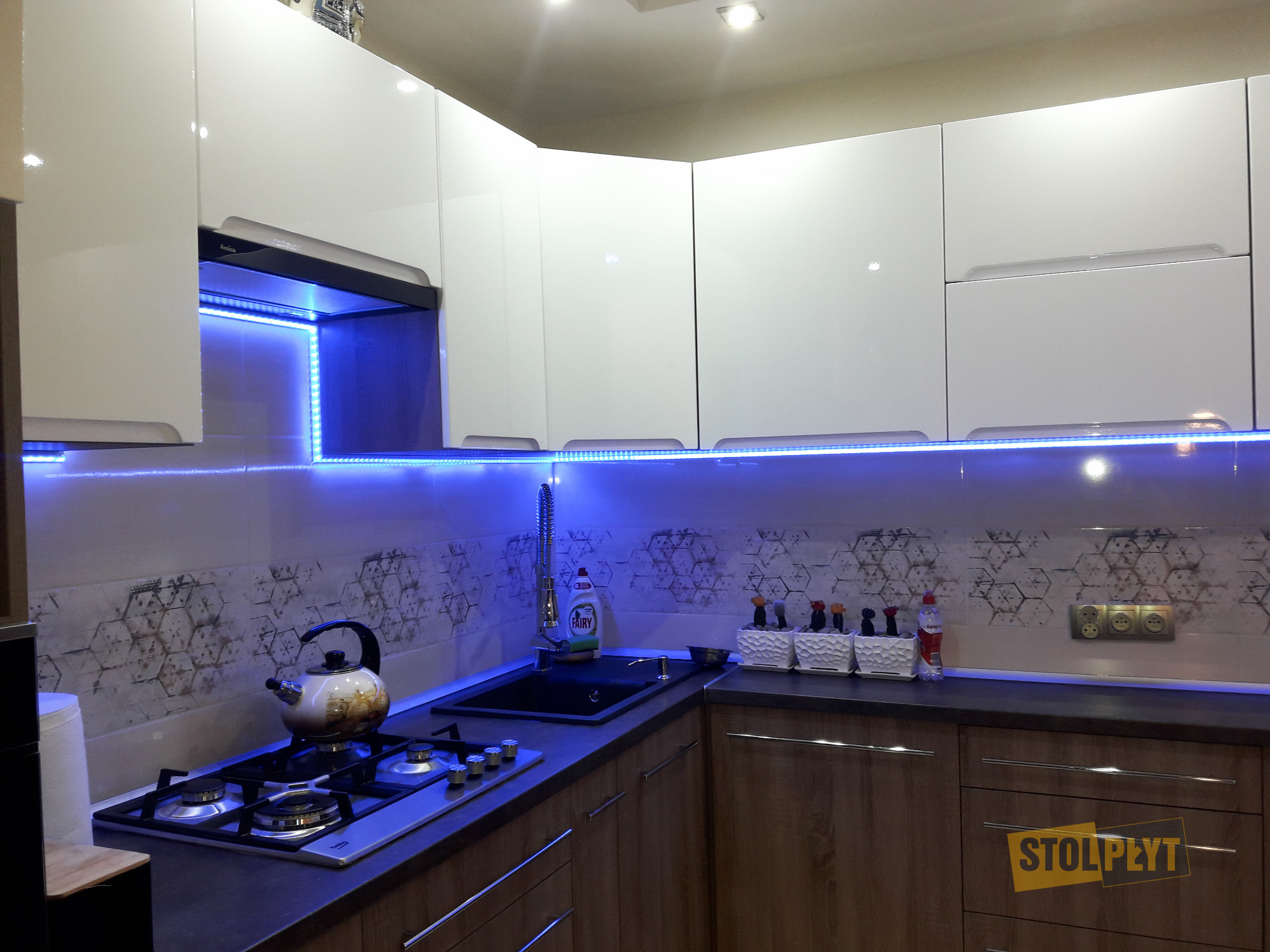 Nowoczesna kuchnia z białymi szafkami górnymi i drewnopodobnymi dolnymi, podświetlona niebieskim LED, z czajnikiem na płycie gazowej i dekoracyjnymi akcentami.