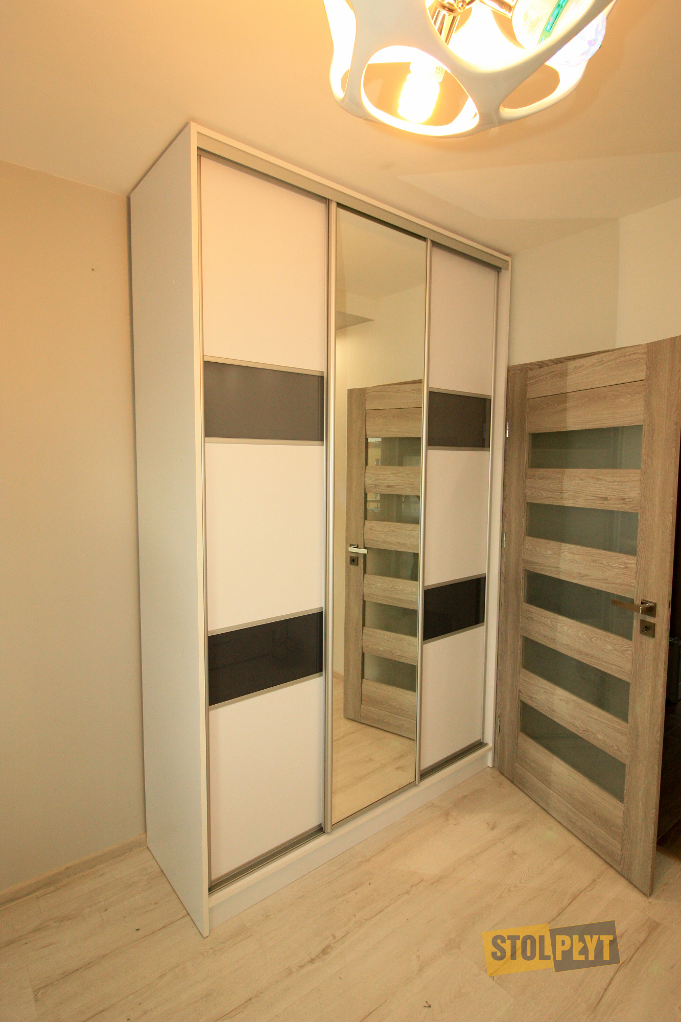 Biała szafa przesuwna z lustrem i ciemnymi akcentami, widoczna w jasnym wnętrzu z drewnianą podłogą i drzwiami, oświetlona nowoczesną lampą sufitową.