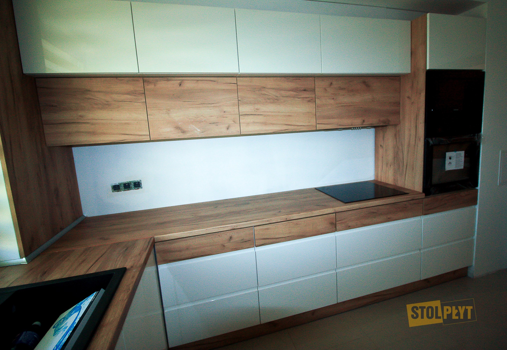 Nowoczesna kuchnia z białymi szafkami dolnymi i drewnopodobnymi górnymi, czarna płyta indukcyjna, widoczne gniazdka elektryczne na ścianie, minimalistyczny design.