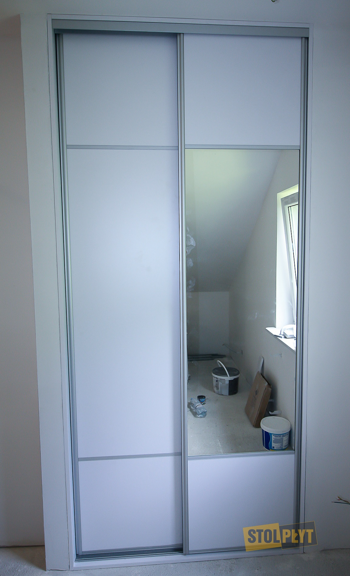 Biała szafa przesuwna z lustrem, widoczne odbicie wnętrza pokoju w trakcie remontu z wiadrami i butelkami.