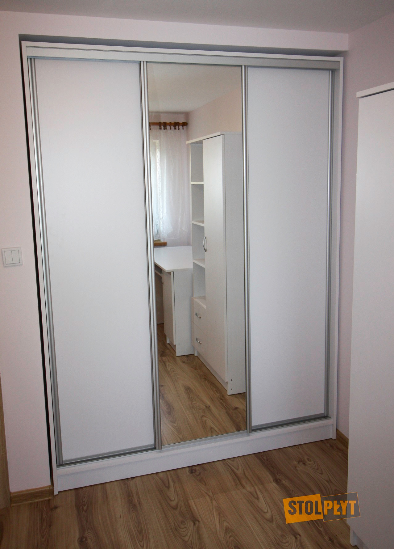 Biała szafa przesuwna z lustrem, widoczny fragment pokoju odbity w lustrze, drewniana podłoga.