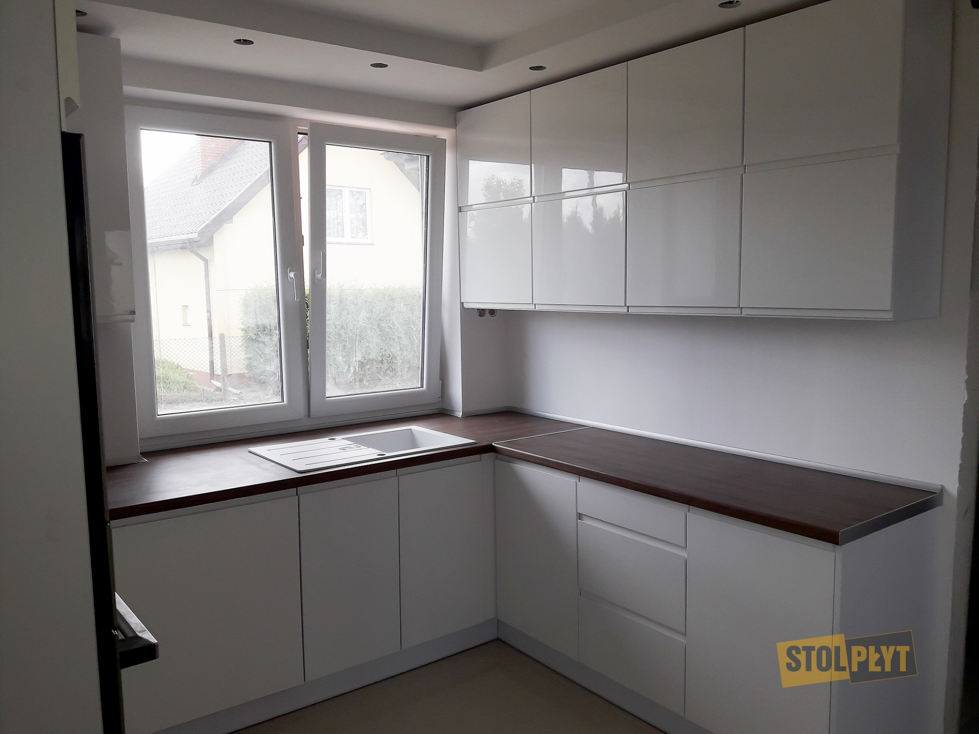 Nowoczesna, biała zabudowa kuchenna z ciemnym blatem, widok na okno z widokiem na dom i ogród, logo firmy Stol-Płyt w prawym dolnym rogu.