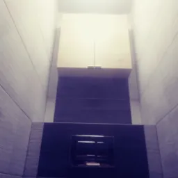 Wykończona łazienka z szarymi płytkami imitującymi drewno, zabudowana szafka nad toaletą z jasnego drewna i czarny panel sterujący spłuczką.