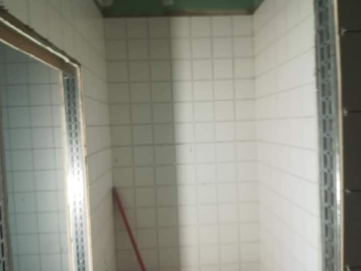 Wnętrze łazienki w trakcie remontu, widoczne białe kwadratowe płytki na ścianach, metalowe profile przy krawędziach, częściowo wykończony otwór drzwiowy, czerwona rurka oparta o ścianę.
