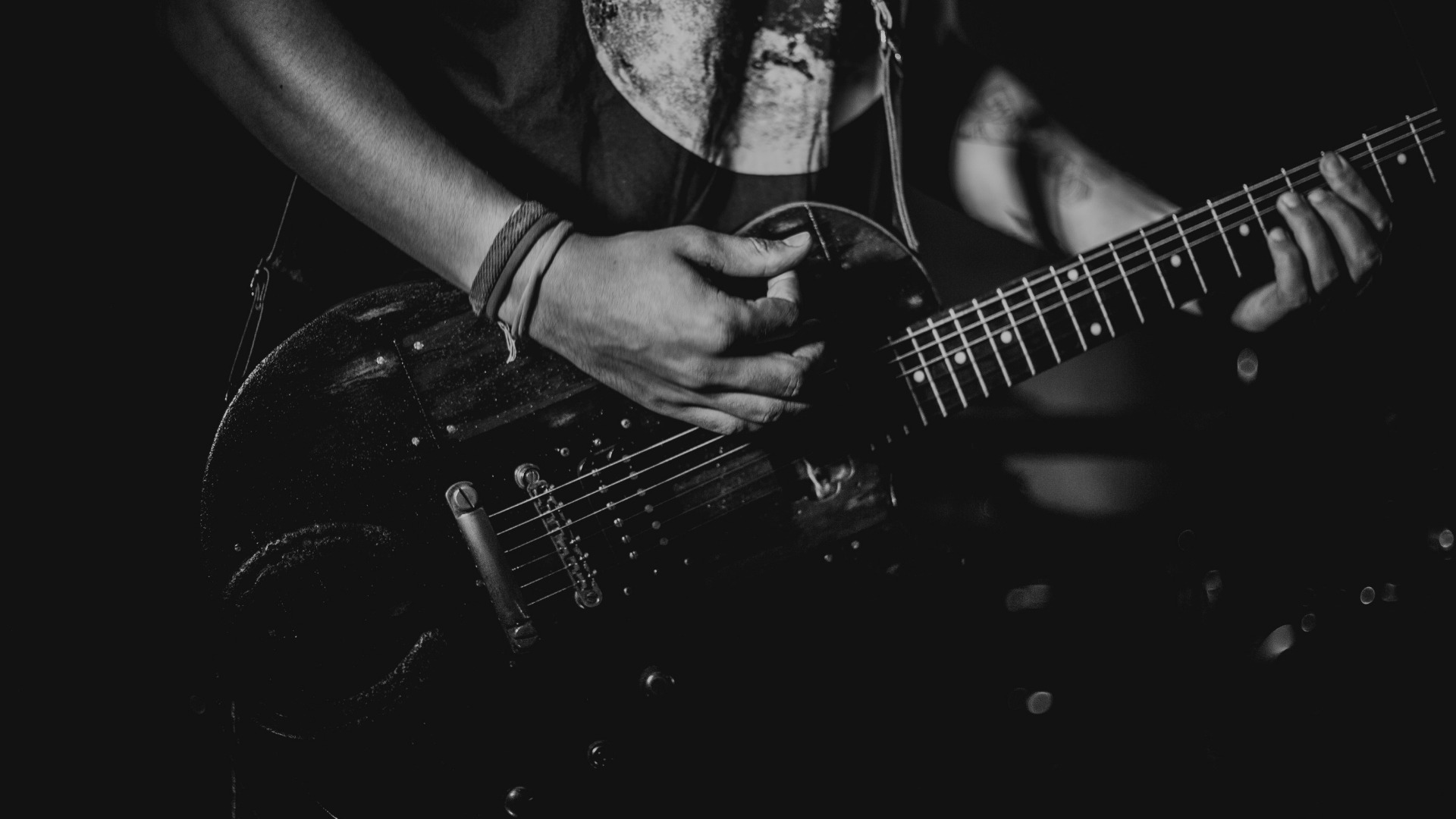 Czarno-białe zdjęcie z bliska przedstawia gitarzystę grającego na elektrycznej gitarze w Zielonkach, widoczne dłonie na gryfie i korpus gitary, ciemne tło z delikatnymi refleksami świetlnymi.