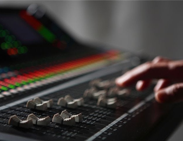 Dłoń delikatnie ustawia suwak na profesjonalnym mikserze audio z podświetlanymi diodami LED w tle, tworząc subtelny efekt bokeh.