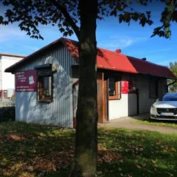 Budynek serwisu AGD z czerwonym dachem i szyldami informacyjnymi, na zewnątrz zaparkowany biały samochód osobowy, widok zza drzewa.