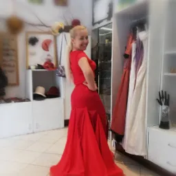 Kobieta w czerwonej sukni wieczorowej, prezentująca strój na tle garderoby z kapeluszami i sukniami w pracowni w Krakowie.