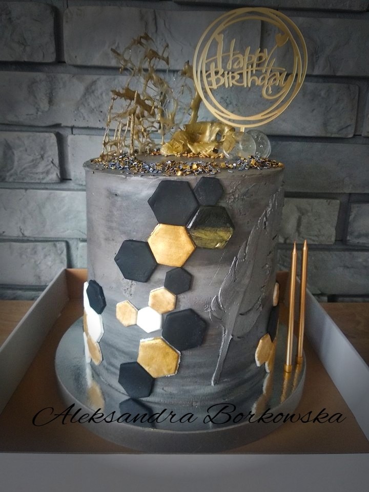 Tort urodzinowy w odcieniach szarości z geometrycznymi dekoracjami w kolorze czarnym i złotym, zwieńczony złotym napisem 'Happy Birthday' i karmelową dekoracją, umieszczony w kartonowym pudełku.