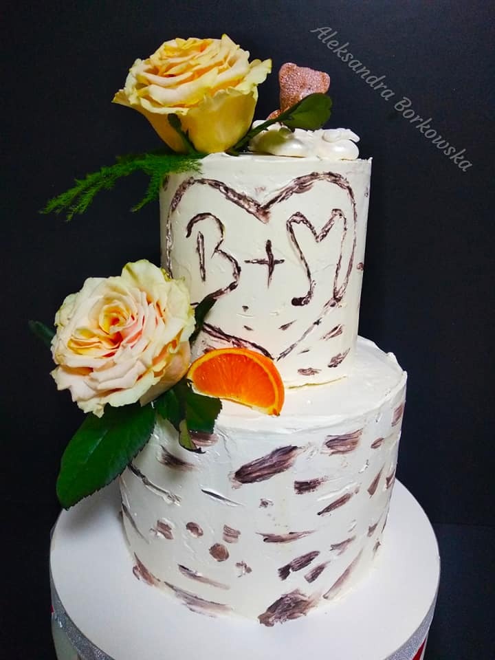 Dwupiętrowy tort weselny w stylu rustykalnym, z kremem imitującym korę brzozy, ozdobiony żółtymi różami, plasterkiem pomarańczy i wyrytym w kremie sercem z inicjałami pary.