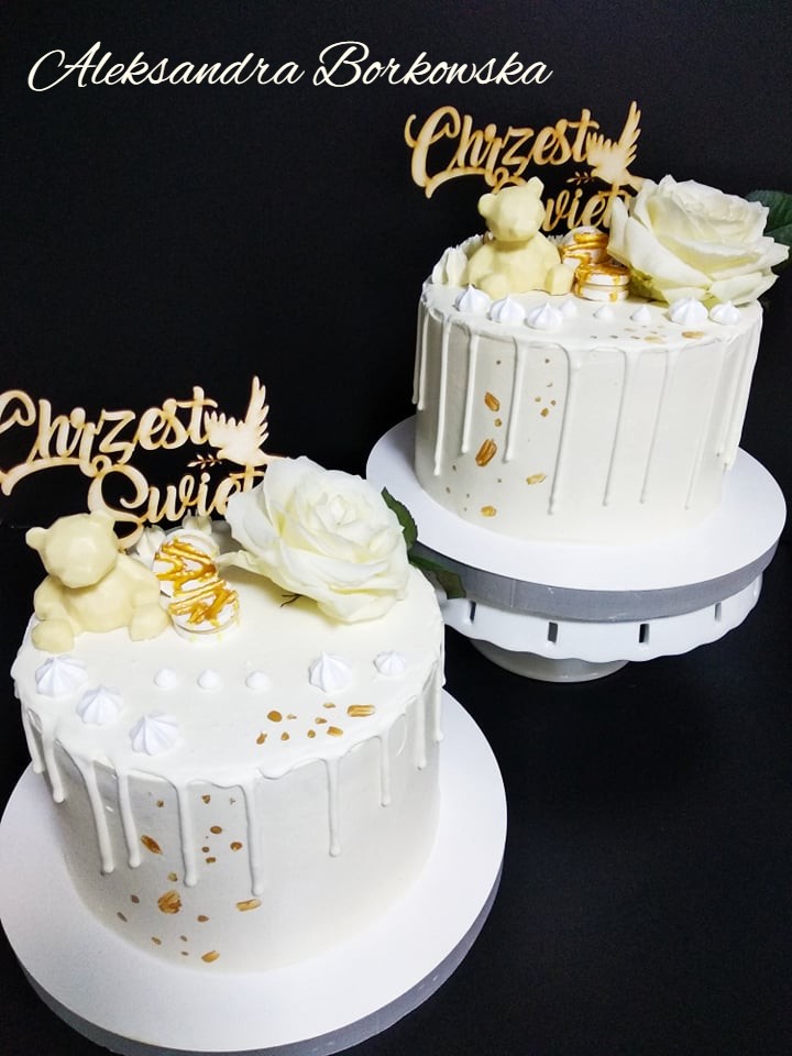 Dwa białe torty z polewą, dekorowane białymi różami, bezikami, złotymi akcentami i drewnianymi napisami 'Chrzest Święty' oraz figurkami białych misiów, ustawione na białych paterach na czarnym tle.