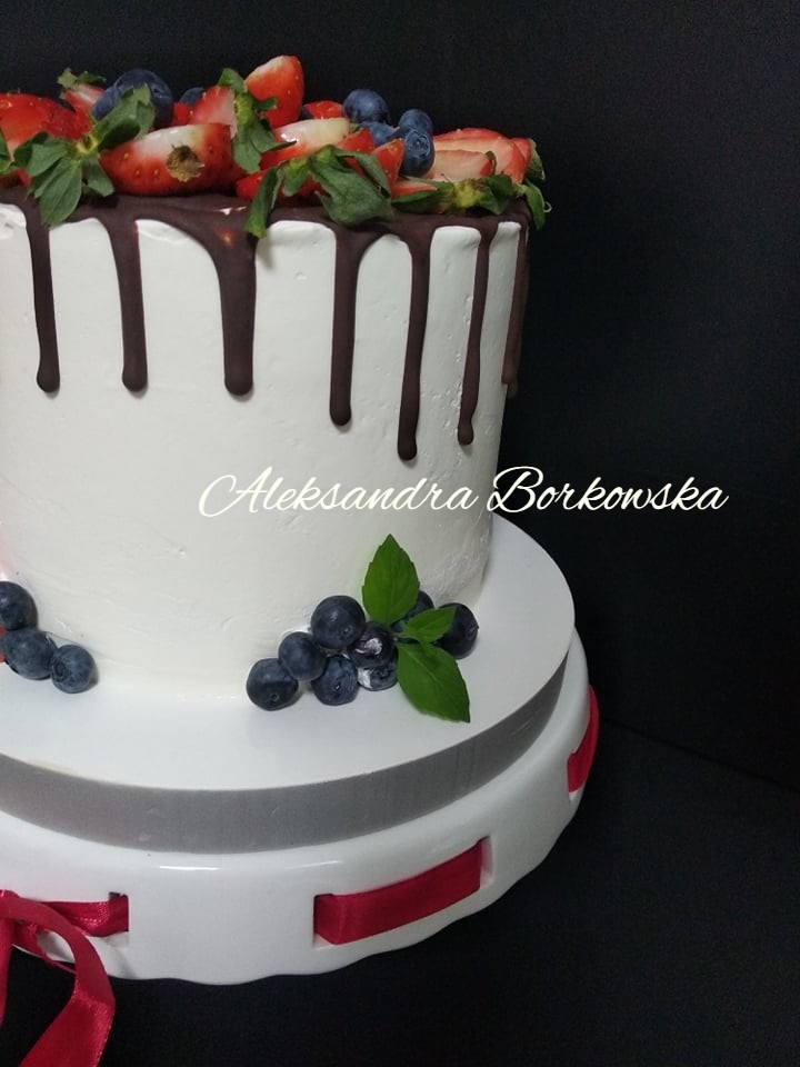Okrągły, biały tort udekorowany polewą czekoladową, świeżymi truskawkami, borówkami i listkami mięty, umieszczony na białej paterze z czerwoną wstążką.
