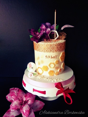 Tort dwupoziomowy na urodziny z dekoracją z fioletowych kwiatów, złotym ornamentem i napisem '59 lat', umieszczony na białym stojaku z różową wstążką na czarnym tle.