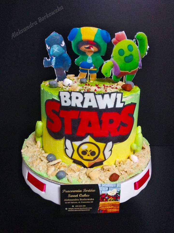 Tort z motywem gry Brawl Stars, udekorowany figurkami postaci Crow, Leon i Spike, z logo gry i posypką imitującą piasek.