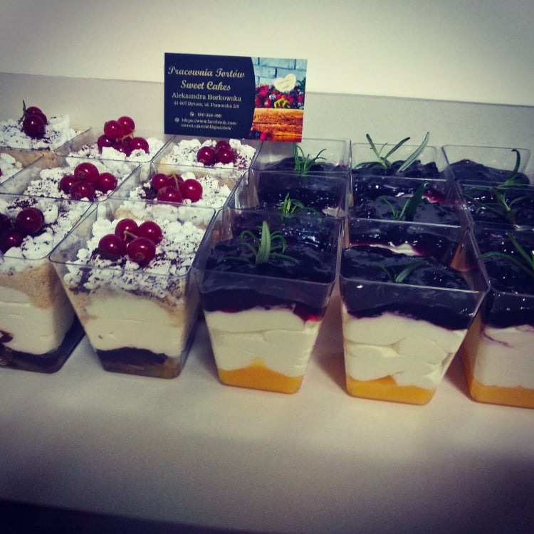 Deserki w plastikowych kubeczkach: po lewej z kremem, posypką i wiśniami, po prawej z galaretką jagodową i gałązką rozmarynu; w tle wizytówka Pracowni Tortów Sweet Cakes Aleksandry Borkowskiej.
